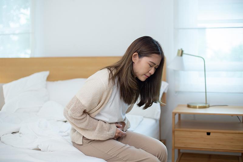 Asian woman sitting on a bed holding her lower abdomen with a pained expression, illustrating abdominal discomfort possibly related to gynaecological issues